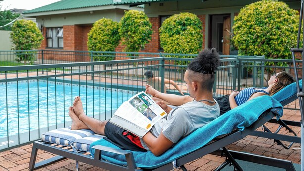A poolside area with lounge chairs, a swimming pool, and a building with a green roof and brick walls. - Premier Guest Lodge (Victoria Falls)