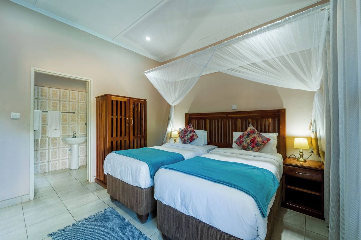 A bedroom with two beds, a wooden wardrobe, a bedside table with a lamp, and a bathroom with a sink and mirror.