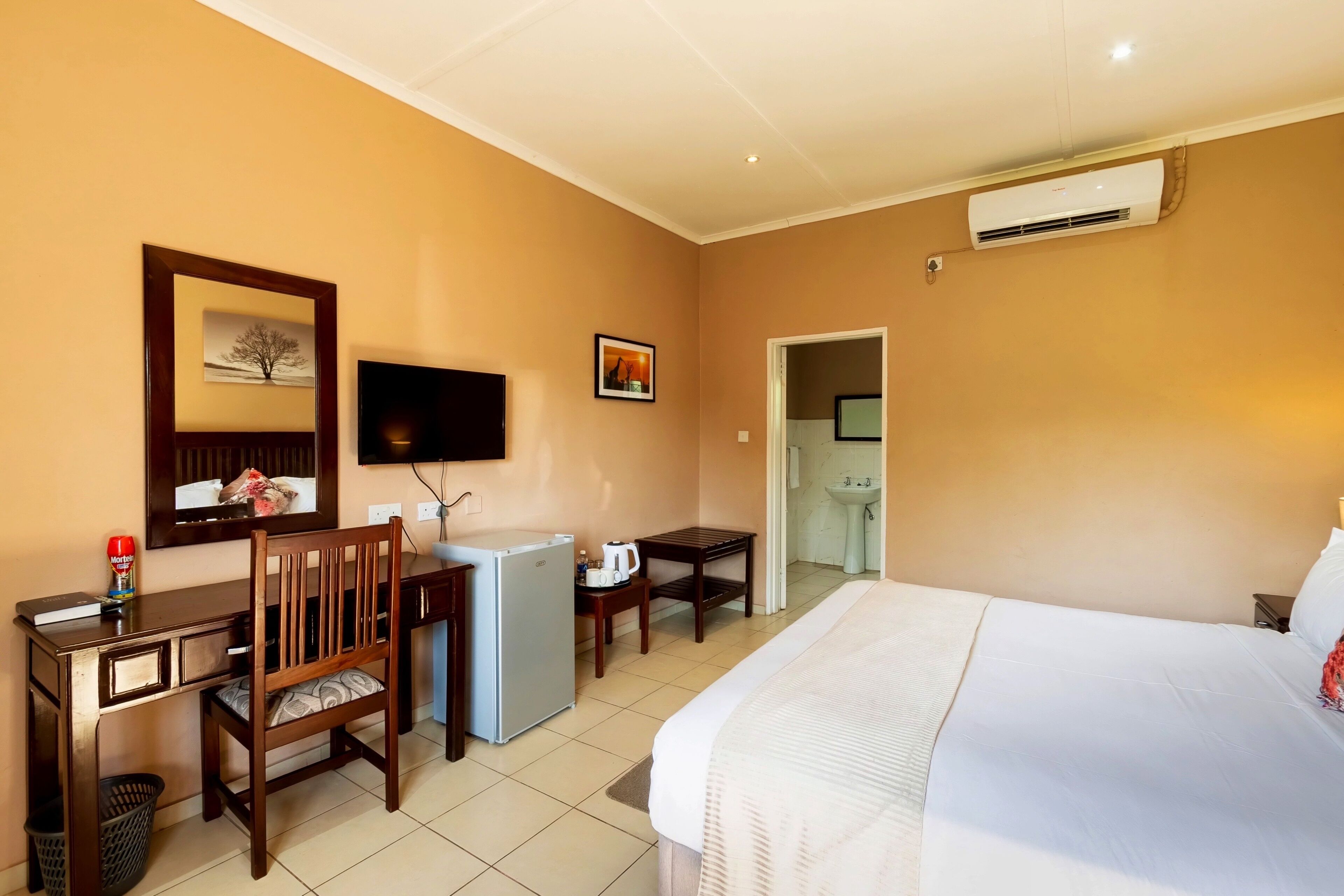 A hotel room with a bed, desk, chair, television, air conditioning unit, and a small kitchenette.