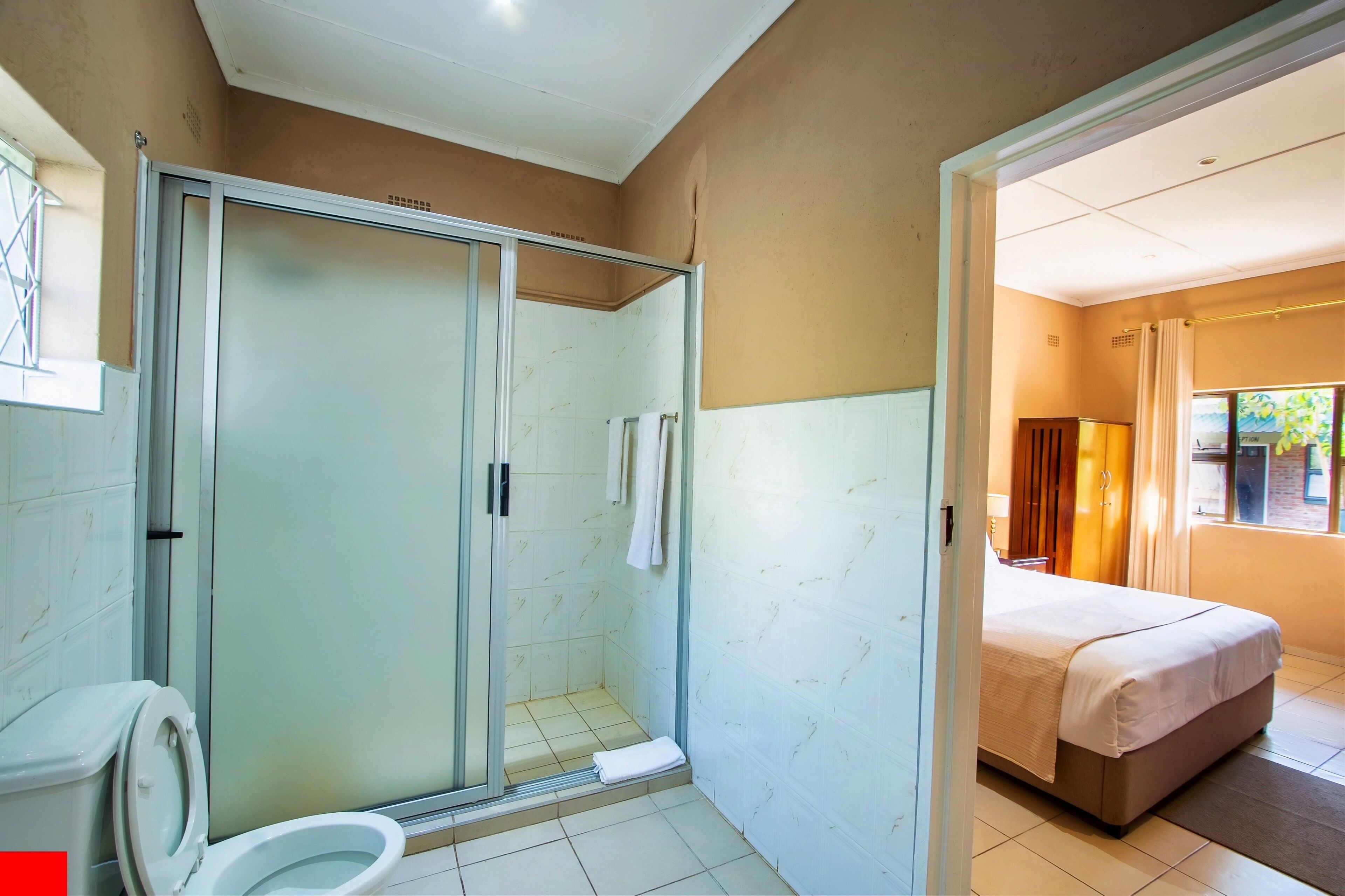 A bathroom with a glass-enclosed shower, a toilet, and a bed in the adjacent room.