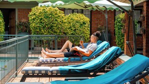 A man relaxing on a sun lounger with a book and a drink, under a green umbrella, with a brick building and greenery in the background. - Premier Guest Lodge (Victoria Falls)