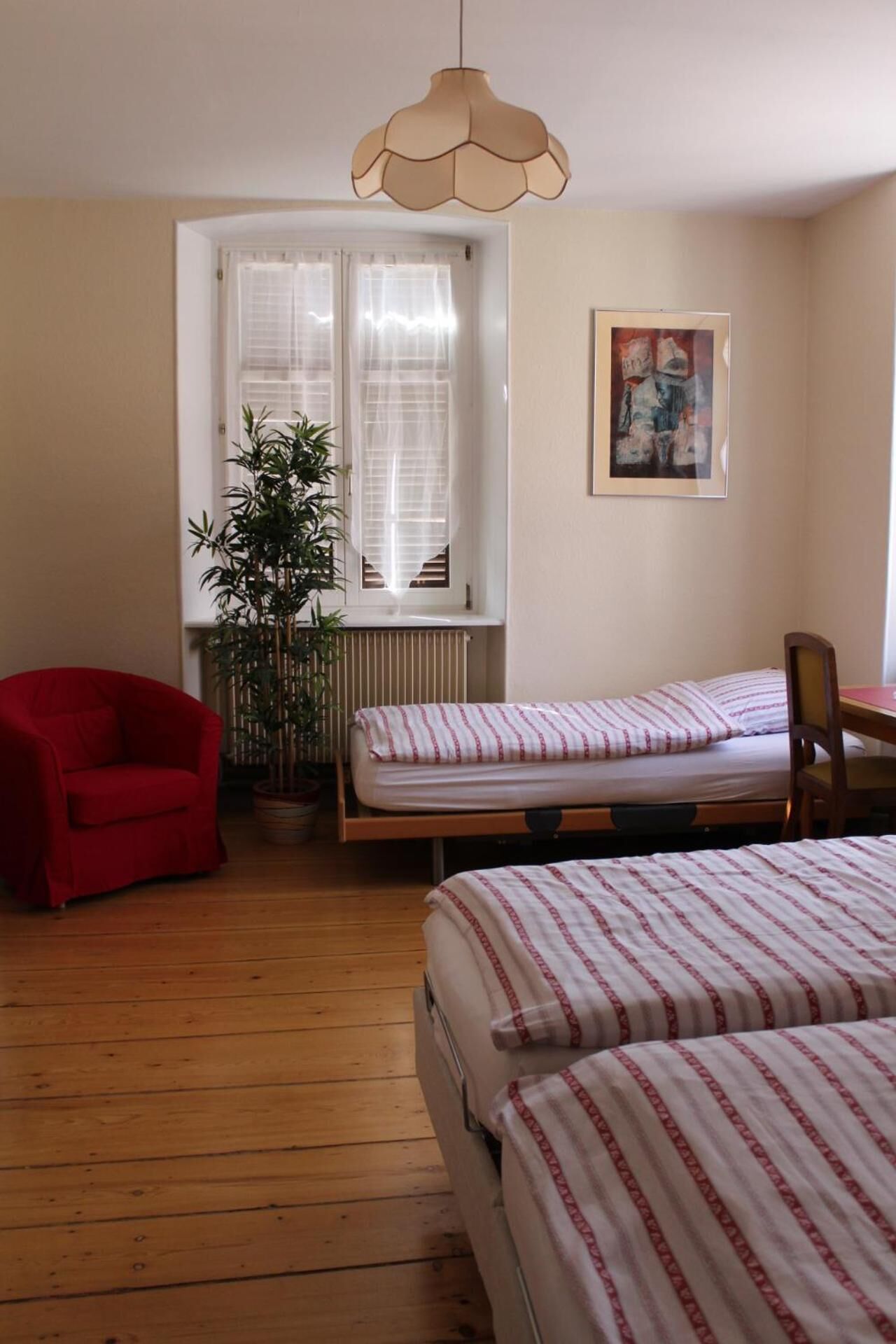 A room with two beds, a red chair, a potted plant, and a window with sheer curtains.