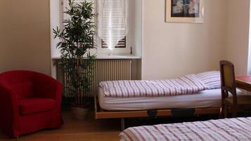 A room with two beds, a red chair, a potted plant, and a window with sheer curtains.