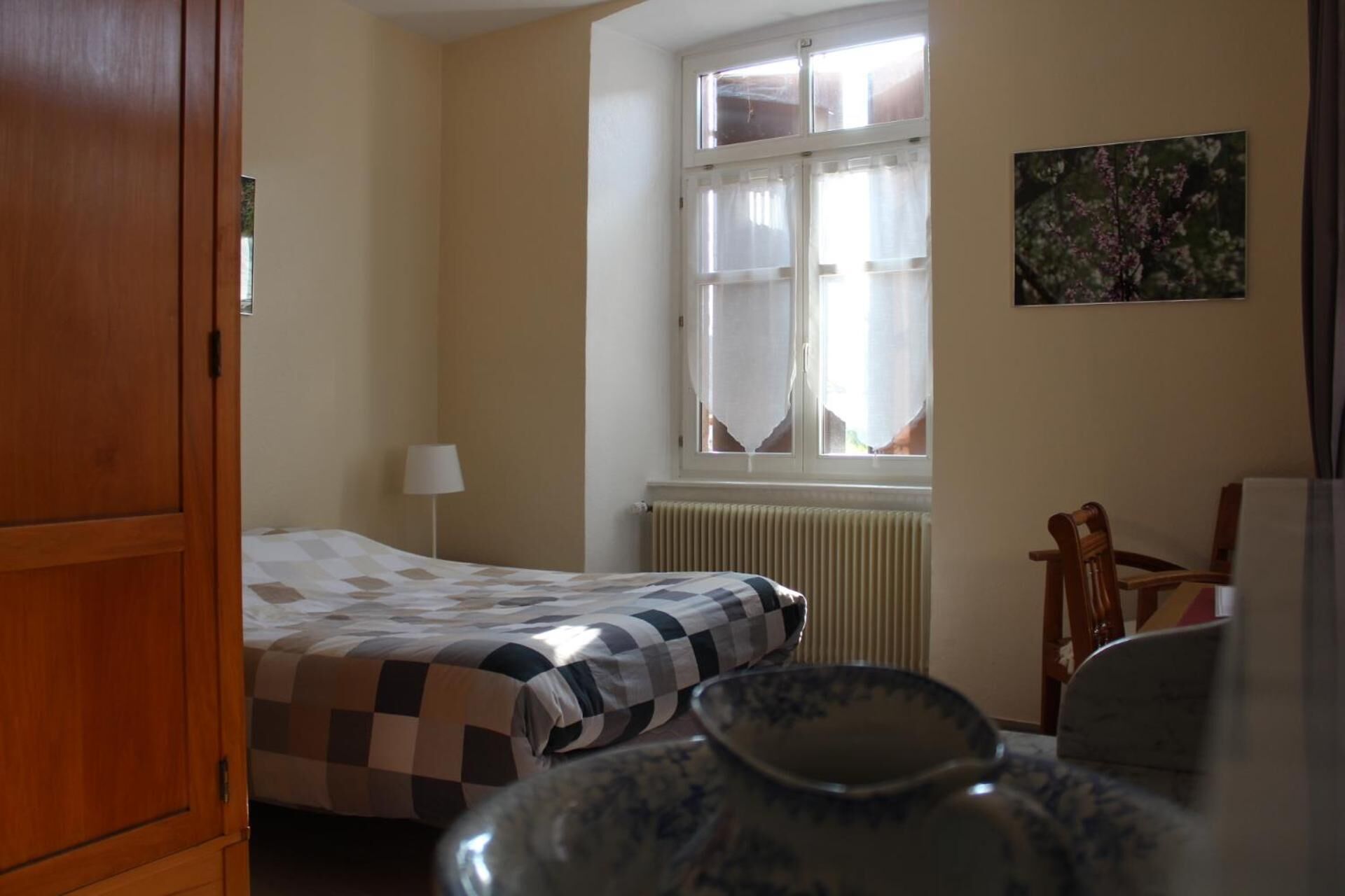 A room with a bed, a radiator, a window with curtains, a chair, and a vase.