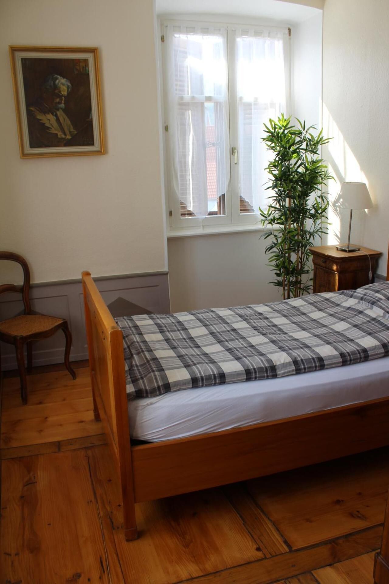 A bedroom with a wooden bed, a chair, a window with curtains, a plant, and a lamp.