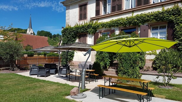 A courtyard with outdoor seating, a green umbrella, and a building with a clock tower in the background. - Ancien Presbytère Albert Schweitzer (Gunsbach)