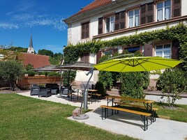 Un patio con mesas al aire libre, un parasol verde y un edificio con una torre de reloj al fondo.