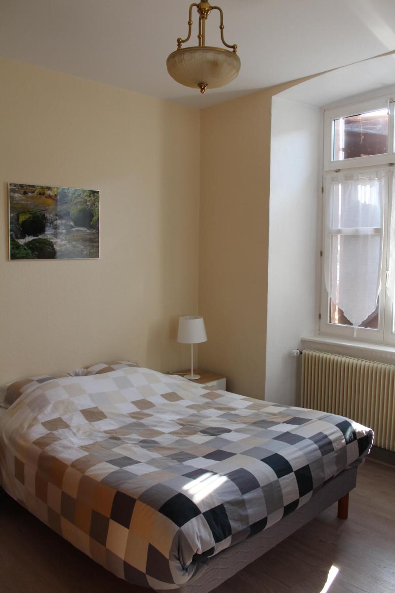 Une chambre à coucher avec un lit, une table de chevet, une lampe, une fenêtre avec des rideaux et un tableau au mur.