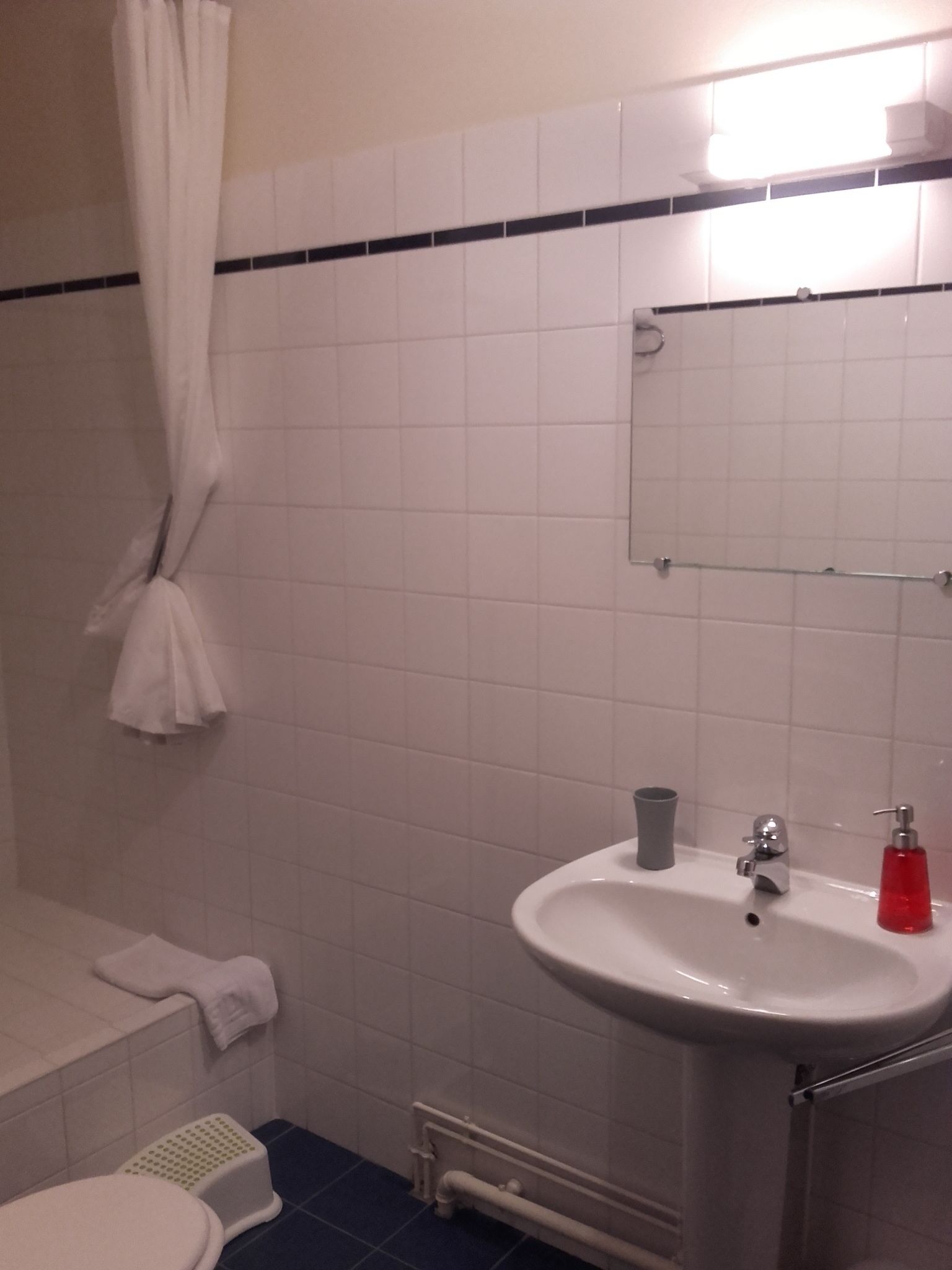 Kylpyhuone, jossa on valkoiset laatat, valkoinen pesuallas, peili, suihkuverho ja pyyhekoukku.