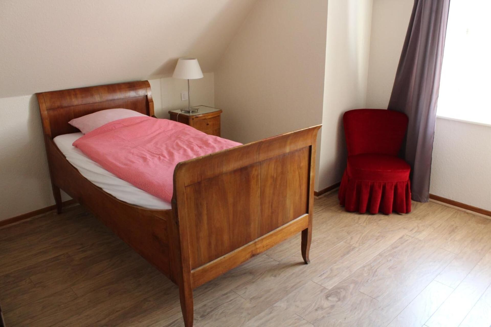 Een houten bed met een roze deken, een rode stoel, een lamp en een raam met gordijnen.