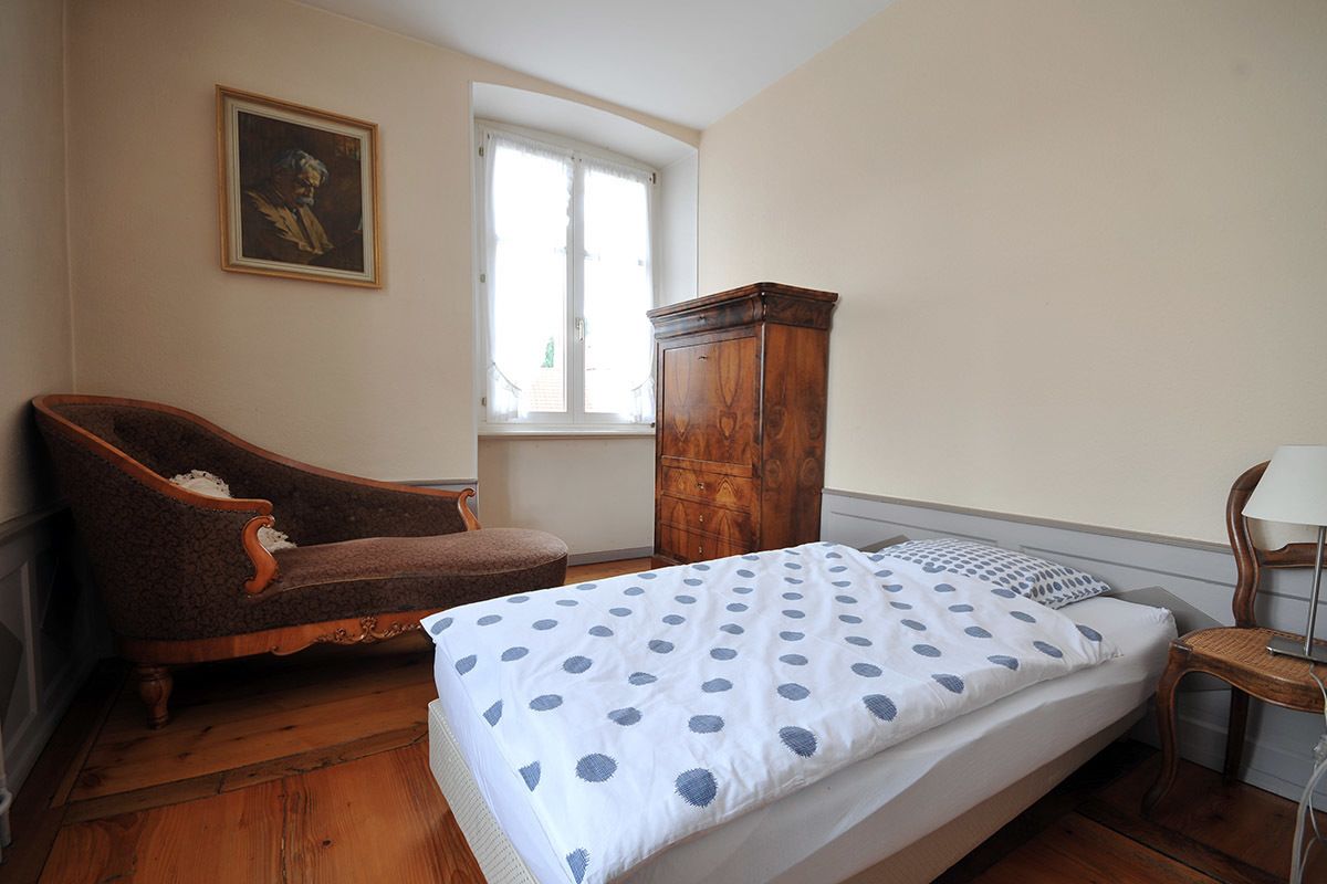 Une chambre à coucher avec un lit, une armoire en bois, un fauteuil marron, une fenêtre et un tableau accroché au mur.