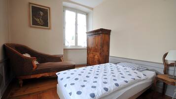 Une chambre à coucher avec un lit, une armoire en bois, un fauteuil marron, une fenêtre et un tableau accroché au mur.
