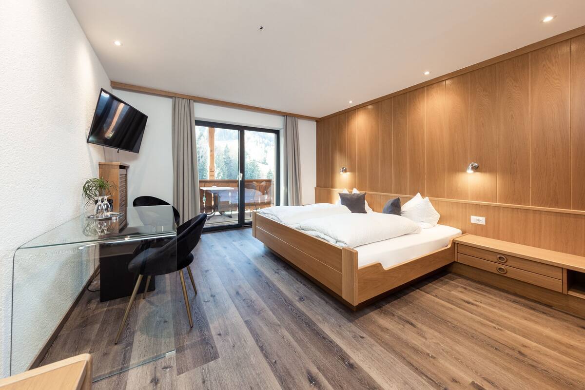 A modern bedroom with a large bed, a desk with a chair, a TV mounted on the wall, and a sliding glass door leading to a balcony.
