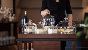 Una zona de bar con un mostrador de madera que exhibe diferentes alimentos bajo campanas de cristal, una persona al fondo y botellas a la izquierda.