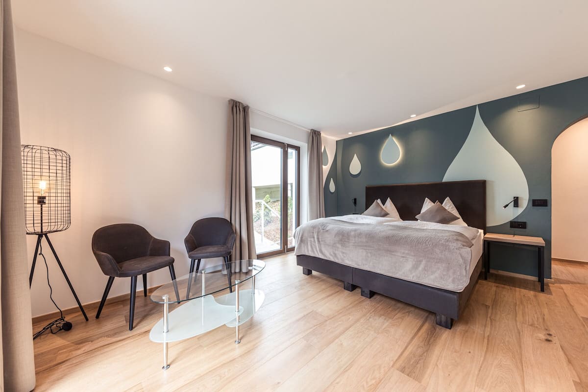 A modern bedroom with a large bed, two chairs, a glass table, and a floor lamp.