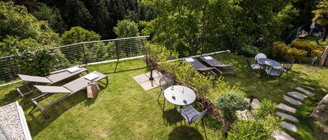 A well-maintained outdoor patio area with lounge chairs, a table, and a barbecue grill, surrounded by lush greenery and a scenic view of a forested valley.