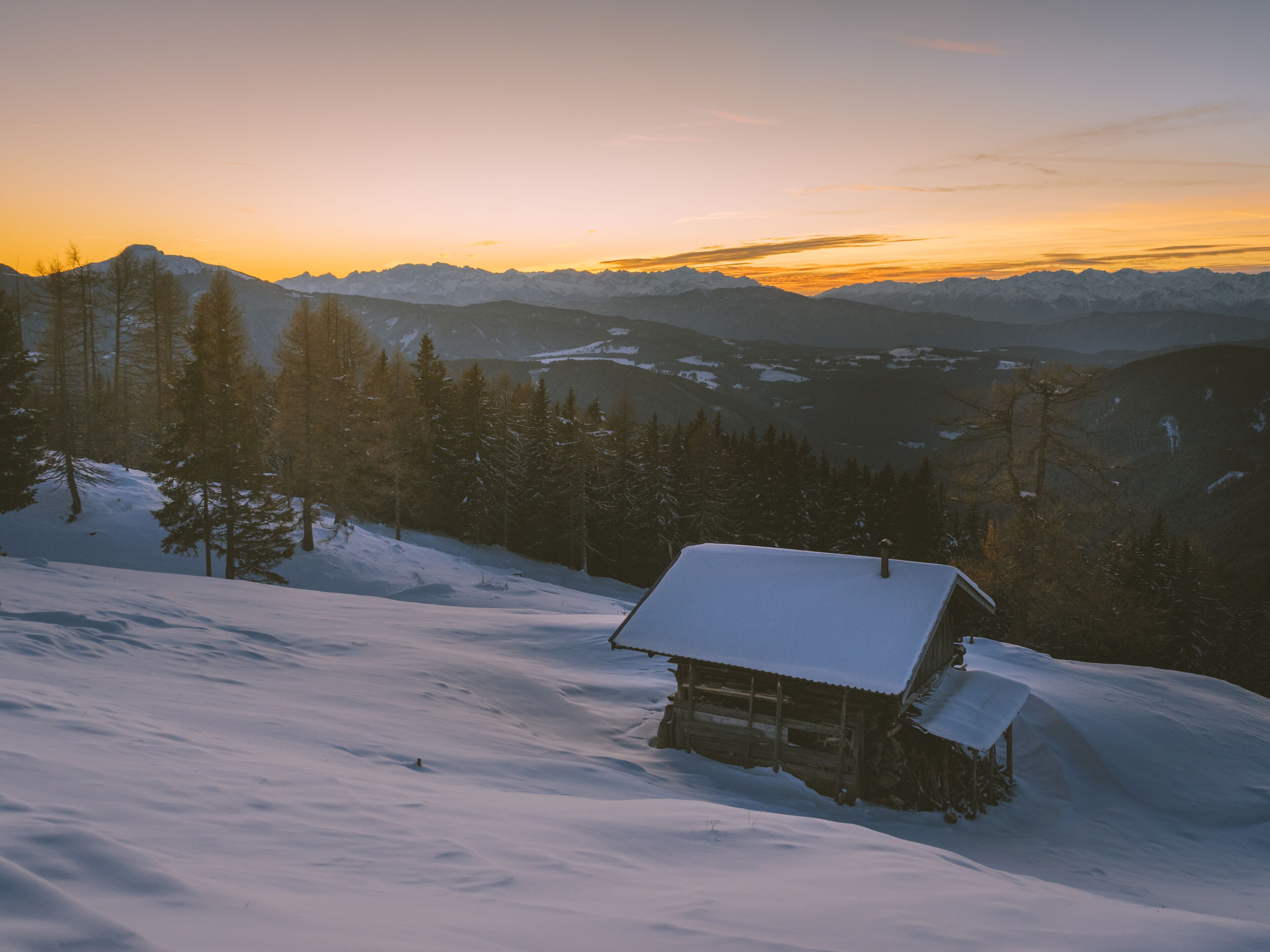Lumihunnun peitt ämä puinen mökki, jonka ympärillä kohoavat lumihuippuiset vuoret ja lumiset puut auringonlaskun aikaan.