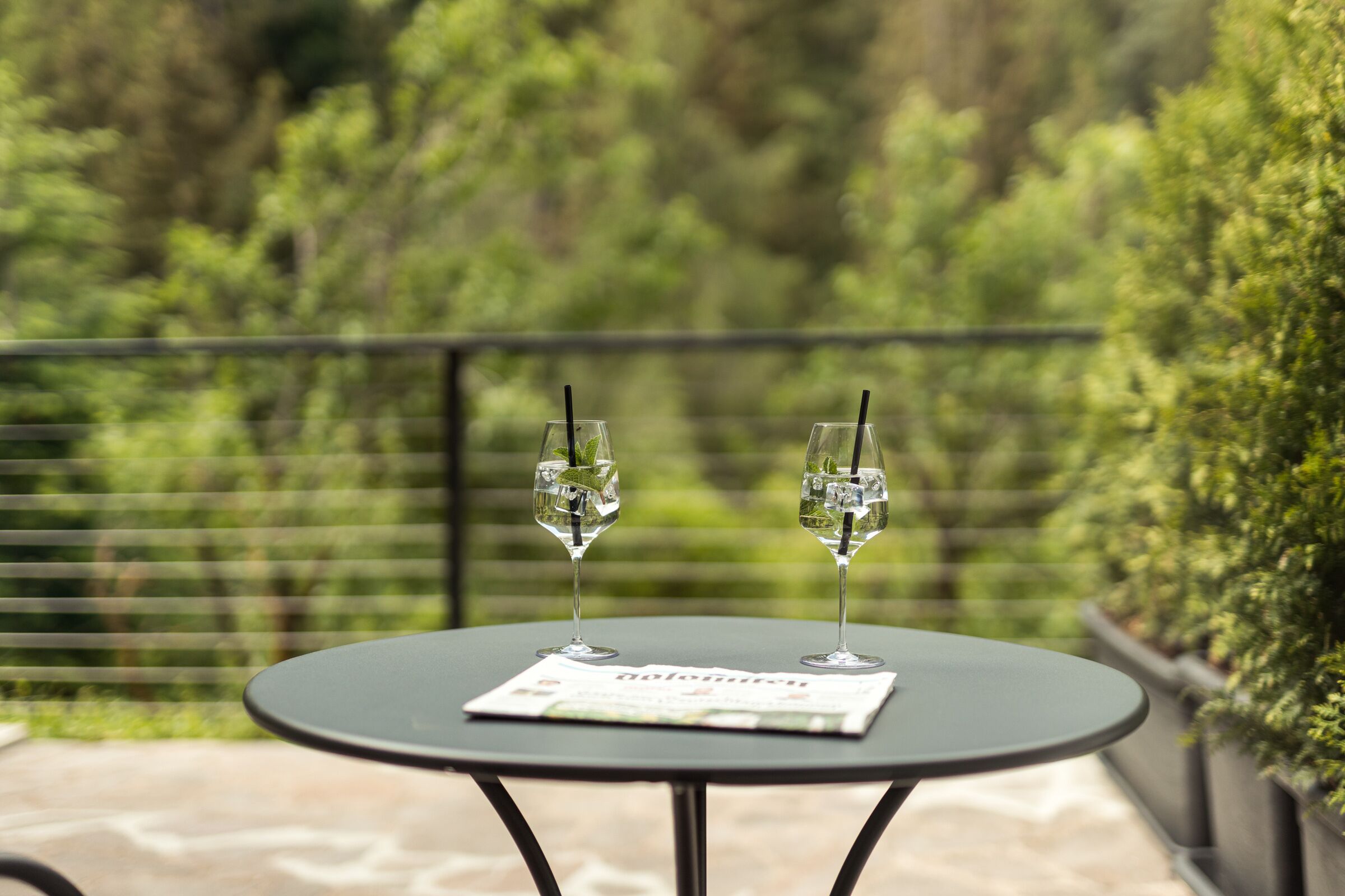 A round table with two glasses of water and a menu on a balcony with a view of greenery.