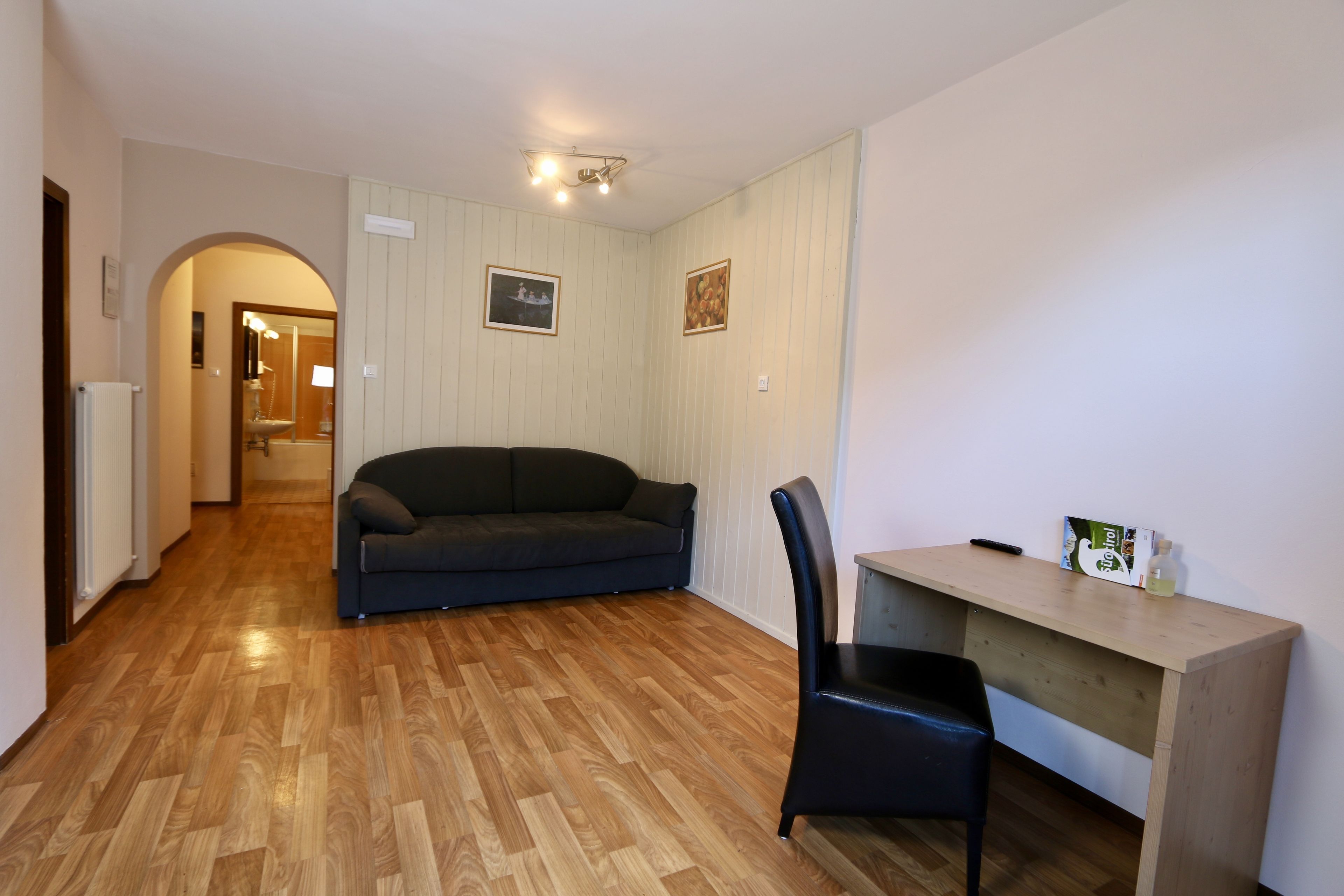 A room with a wooden floor, a dark sofa, a wooden desk, and a black chair.