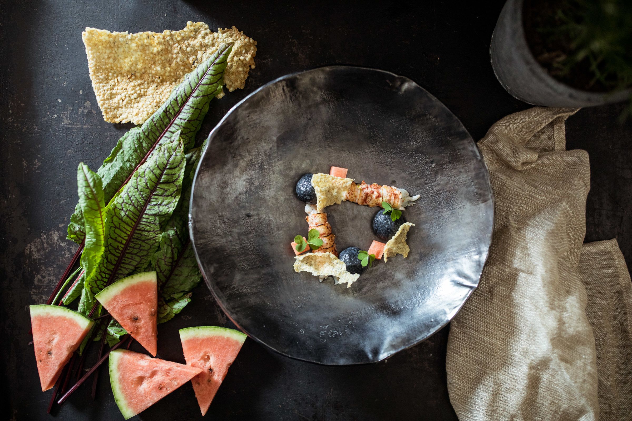 A black plate with food items, a piece of bread, and a cloth napkin on a dark surface.