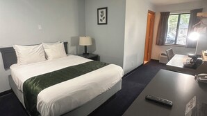 A hotel room with a bed, bedside table, lamp, and a desk with a remote control. - Pictured Rocks Inn & Suites (Munising)