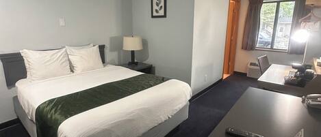 A hotel room with a bed, bedside table, lamp, and a desk with a remote control.