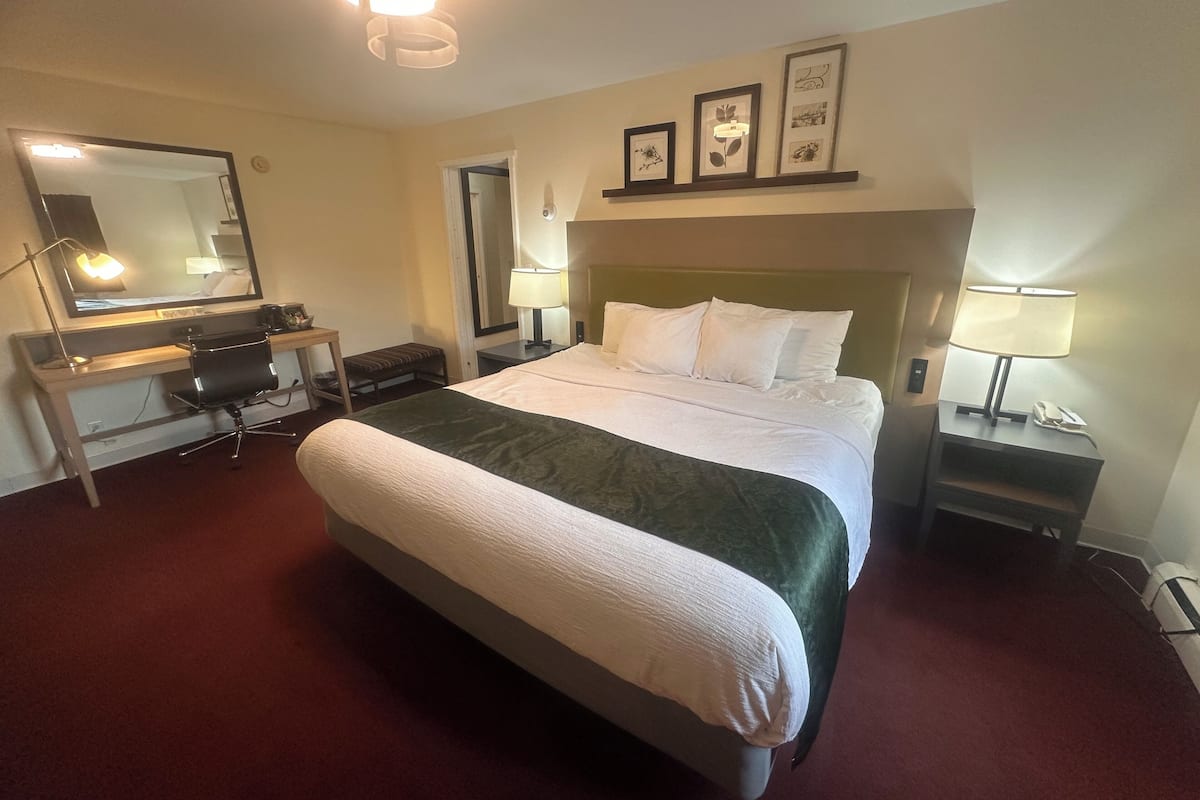 A hotel room with a large bed, a desk with a chair, a mirror, a bedside table with a lamp, and framed pictures on the wall.