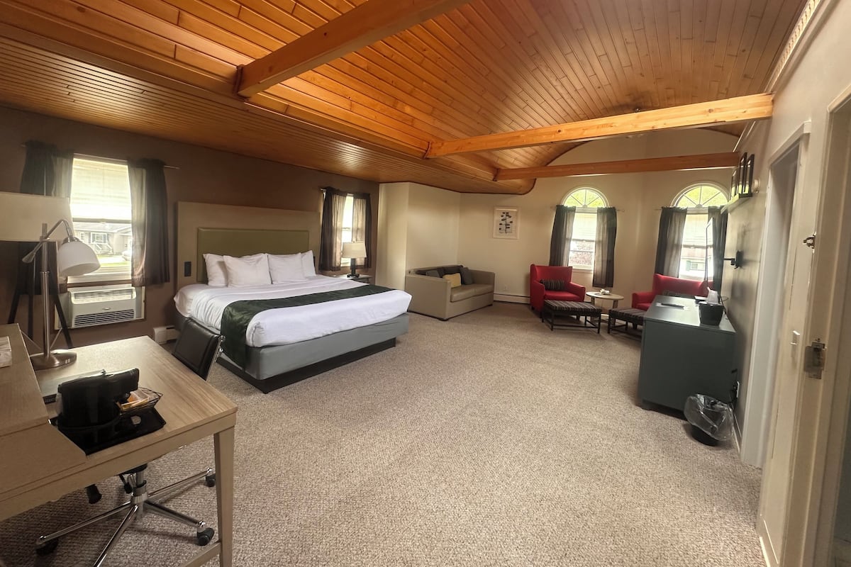 A hotel room with a large bed, a desk, a red armchair, a small table, and a window with curtains.