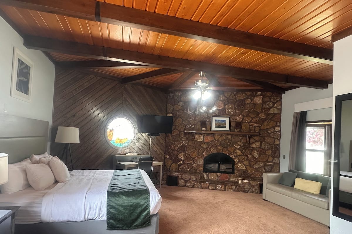 A bedroom with a bed, a desk, a stone fireplace, a television, and a window.