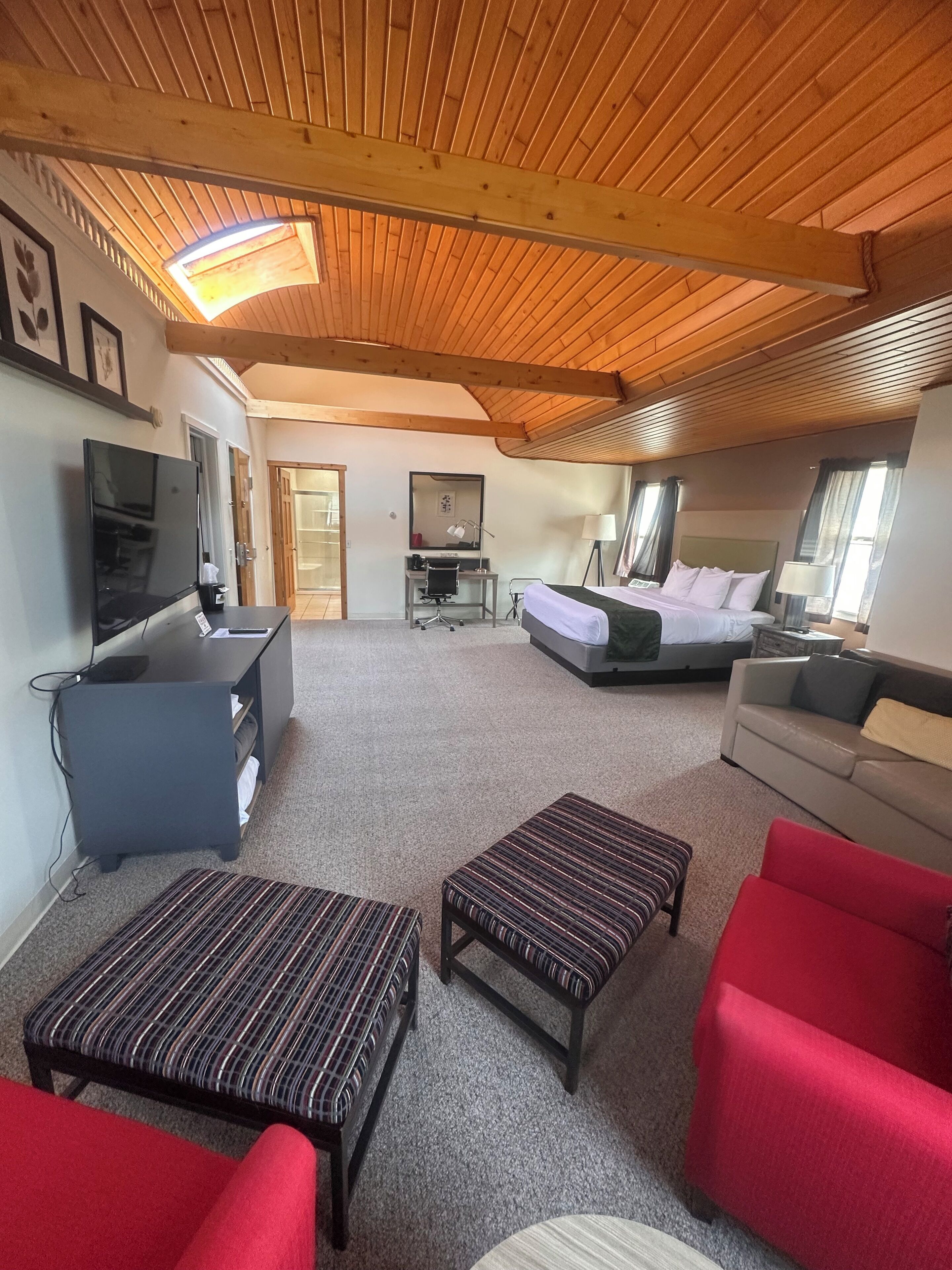 A cozy hotel room with a wooden ceiling, a bed, a desk with a chair, a TV, and a sofa.
