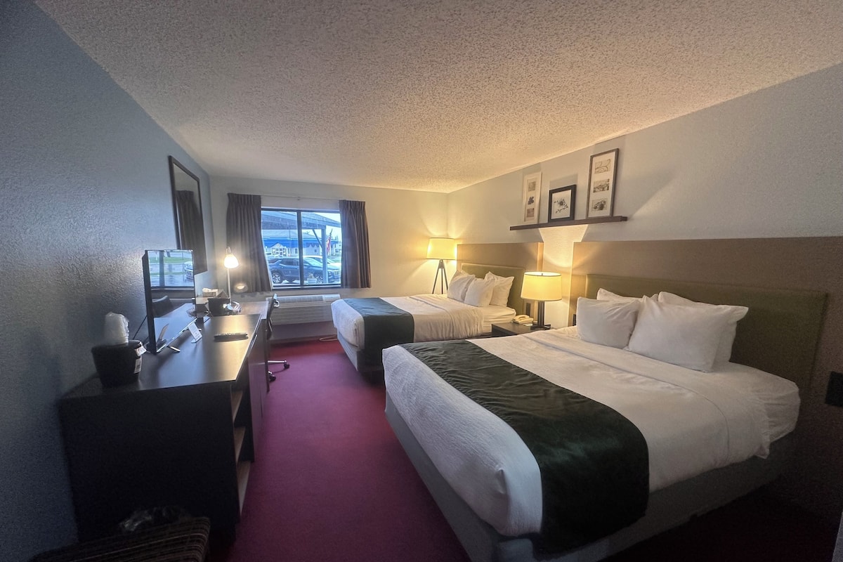 A hotel room with two beds, a desk, a chair, a lamp, and a window with a view of a parking lot.
