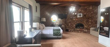 A cozy bedroom with a stone fireplace, a bed with a green blanket, a desk with a lamp, and a large mirror.