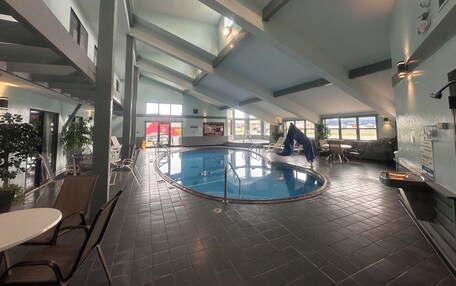 An indoor swimming pool with a tiled floor, a pool ladder, and a glass barrier. There are lounge chairs and tables around the pool area.