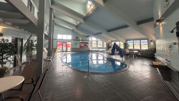 An indoor swimming pool with a tiled floor, a pool ladder, and a glass barrier. There are lounge chairs and tables around the pool area. - Pictured Rocks Inn & Suites (Munising)