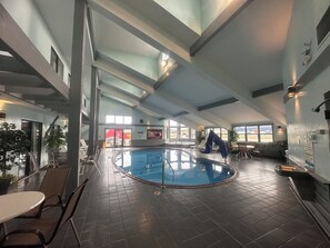 An indoor swimming pool with a tiled floor, a pool ladder, and a glass barrier. There are lounge chairs and tables around the pool area. - Pictured Rocks Inn & Suites (Munising)