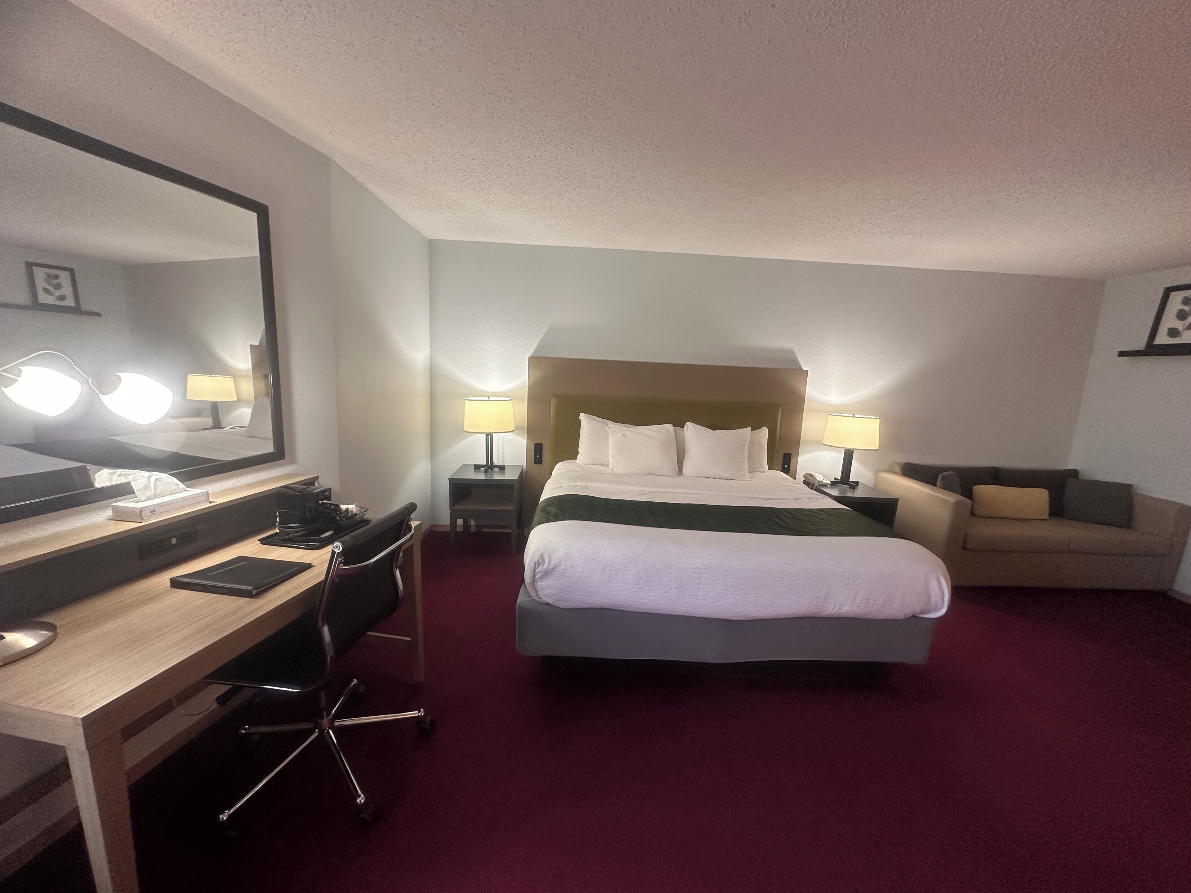 A hotel room with a large bed, a desk with a chair, a desk lamp, a chair, and a bedside table with a lamp.