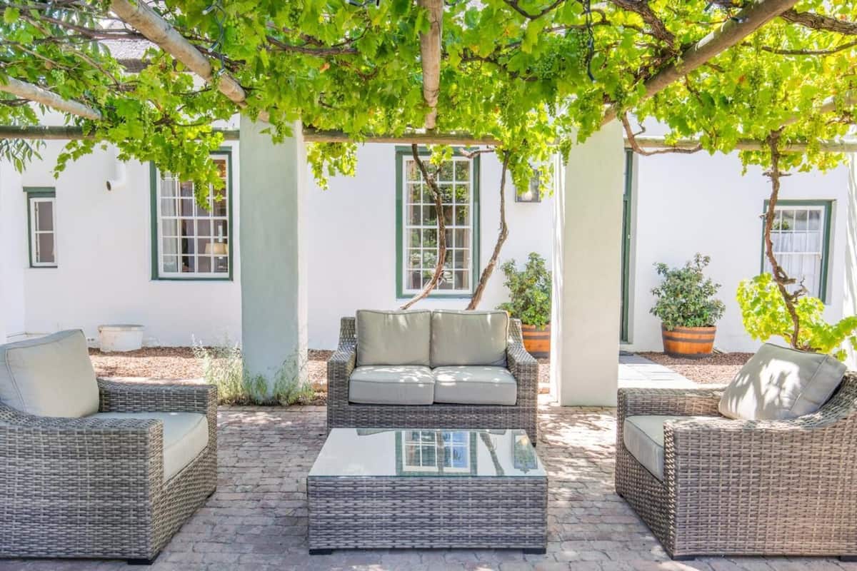 An outdoor seating area with wicker furniture and a glass-top table, surrounded by potted plants and a white building with green window frames.