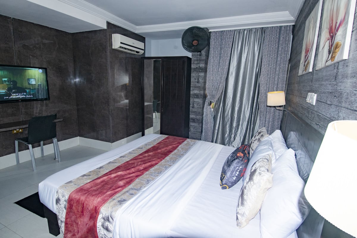 A hotel room with a large bed, a flat-screen TV mounted on the wall, a desk with a chair, and an air conditioning unit.