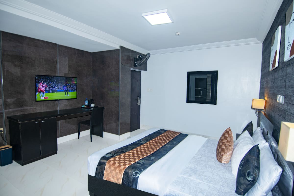 A modern hotel room with a large bed, a flat-screen TV mounted on the wall, a desk, and a dark-colored wardrobe.