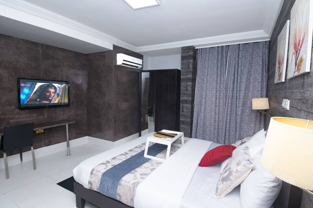 A modern hotel room with a bed, a TV, a desk, and an air conditioning unit.