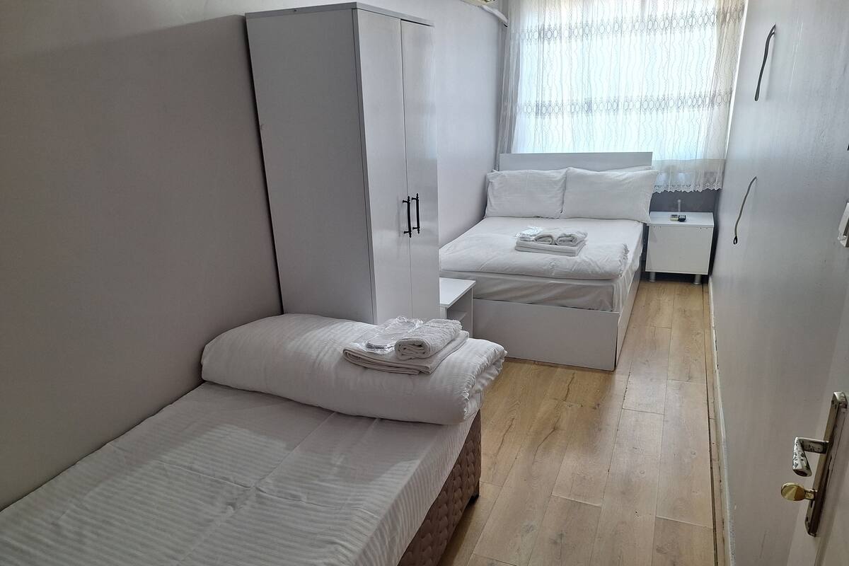 A compact hotel room with two single beds, a wardrobe, a small bedside table, and a window with sheer curtains.
