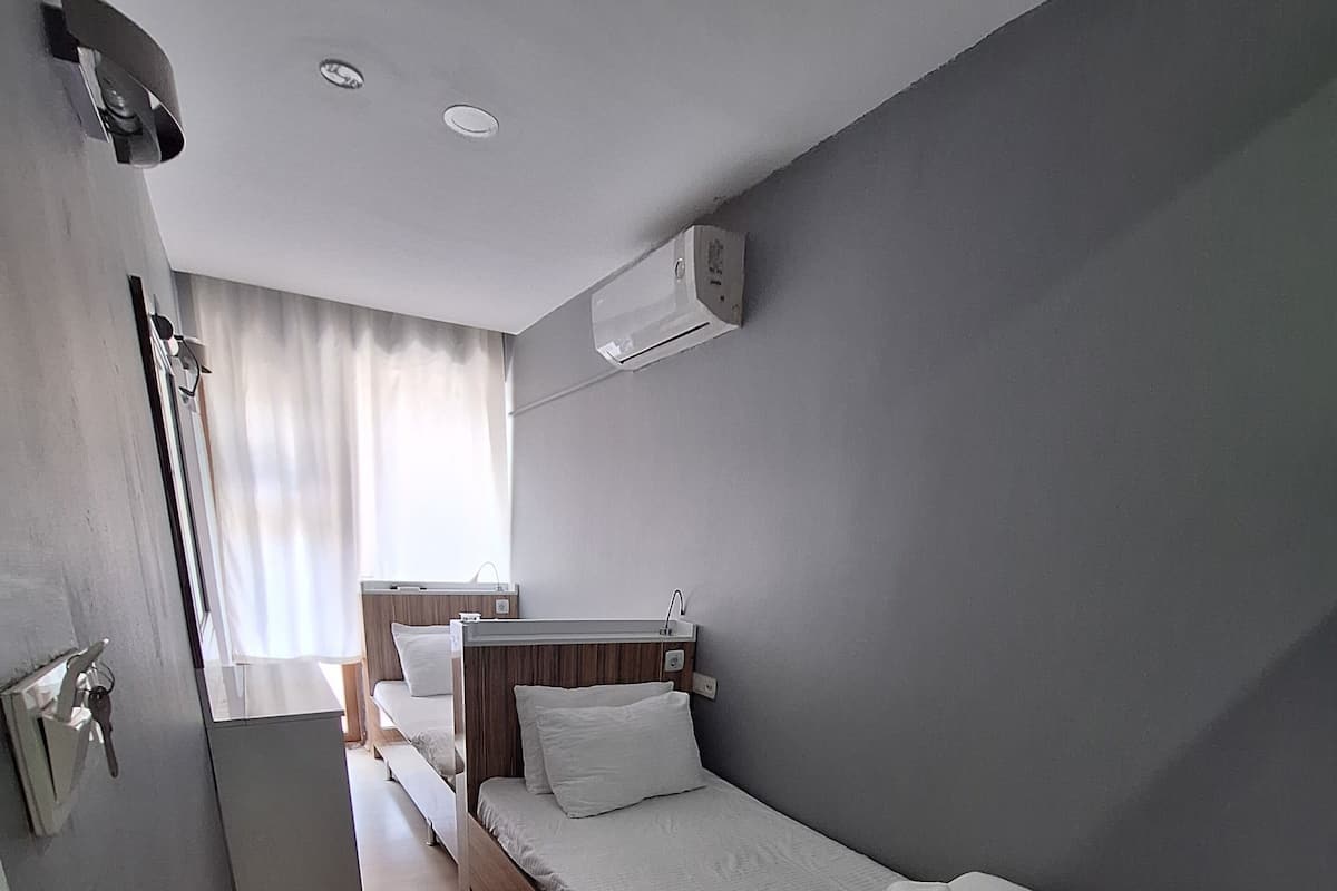 A small, single-bed room with a wooden headboard, a bedside table, a window with curtains, and an air conditioning unit on the wall.