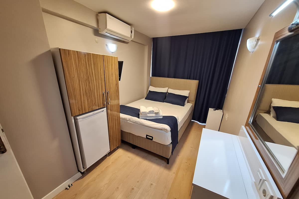 A compact hotel room with a bed, a small desk, a wardrobe, a mini-fridge, and an air conditioning unit.