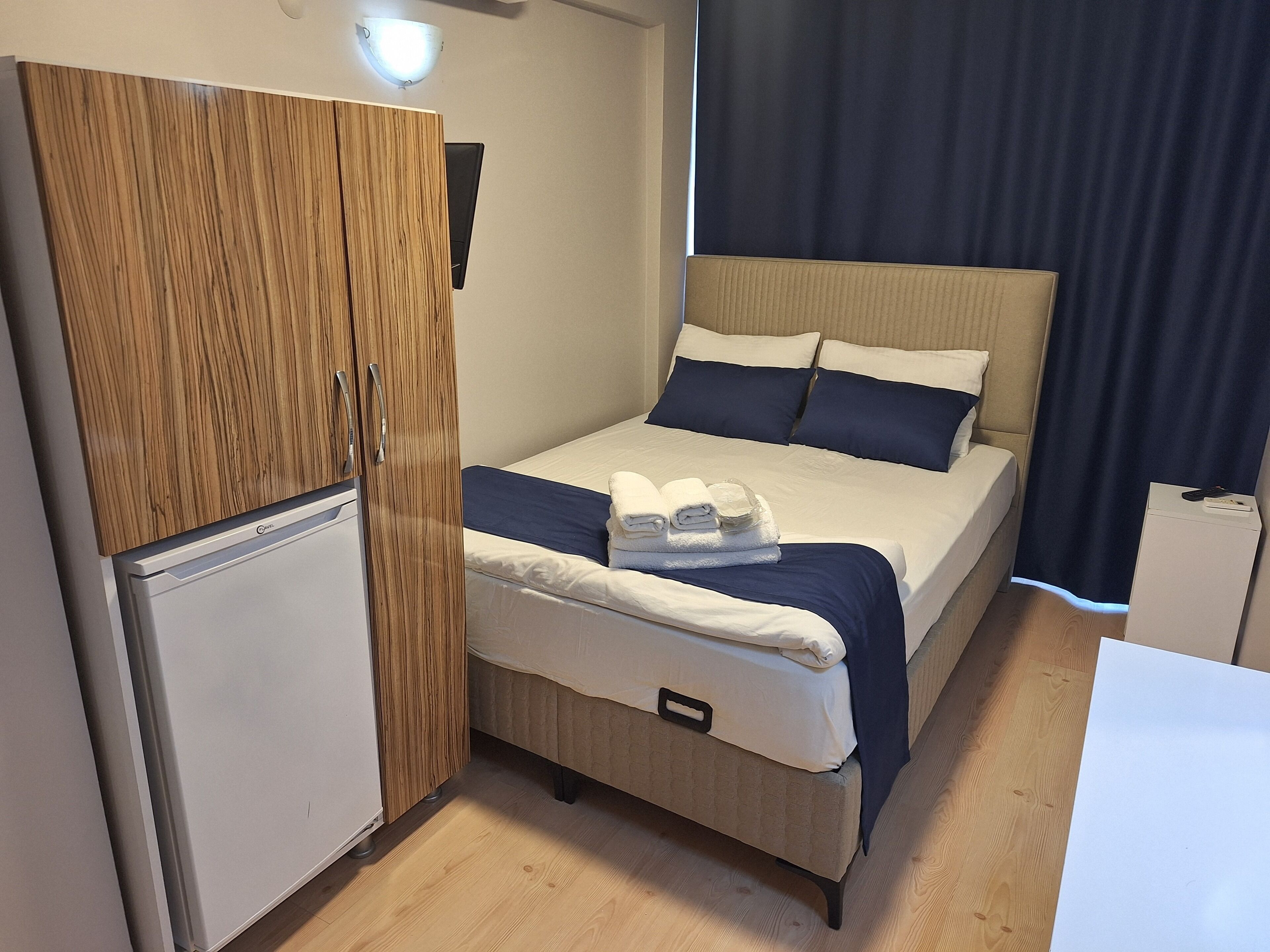 Quarto compacto com cama, guarda-roupa, pequeno frigorífico e mesa de cabeceira.