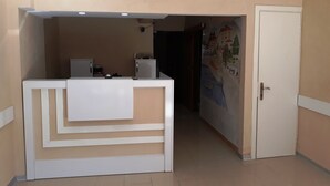 A reception area with a white reception desk, a door to the right, and a tiled wall with a mural. - Dilmac Hotel (Gallipoli)