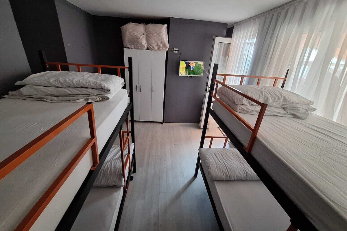 A dormitory room with bunk beds, a wardrobe, a television, and a window with curtains.