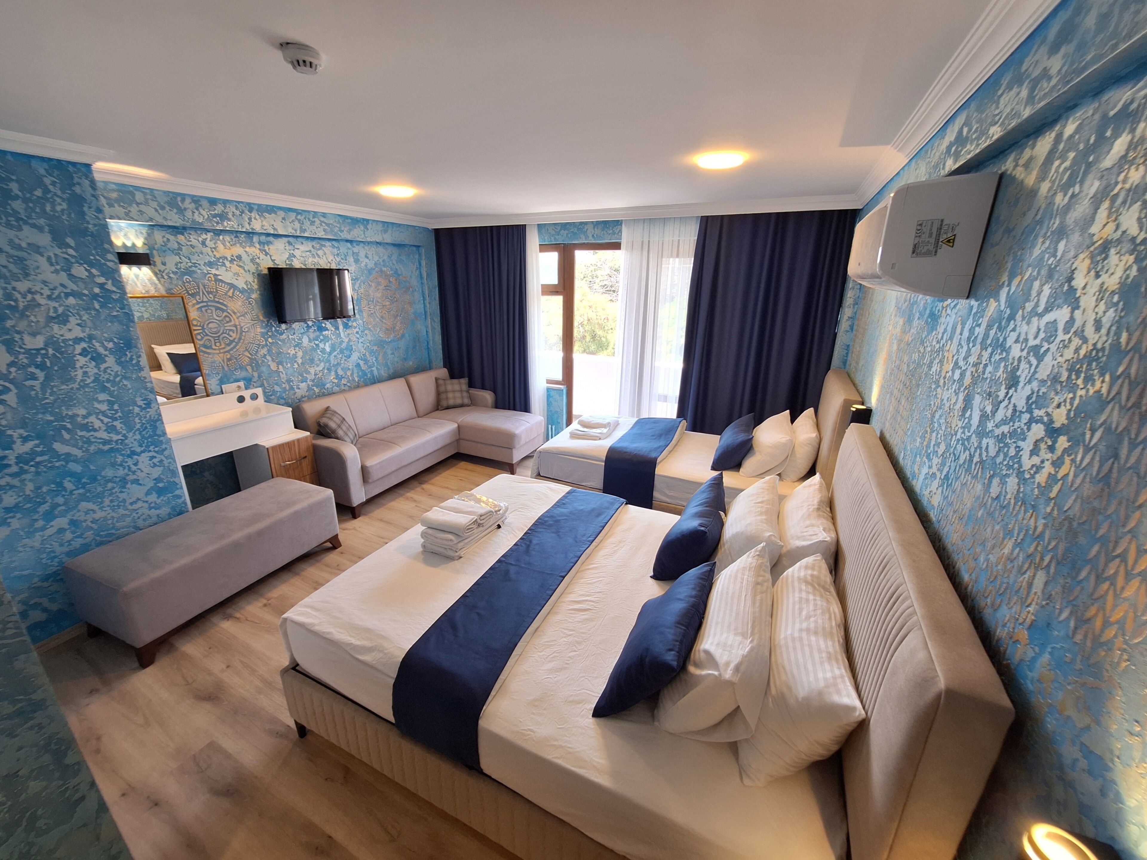 Quarto de hotel com sofá, duas camas, uma mesa de trabalho e uma janela grande.