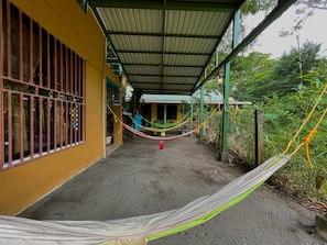 A covered walkway with a hammock, a building with barred windows, and a green roof.