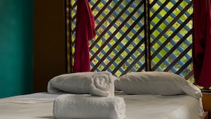 Cama bem arrumada com roupa de cama branca e toalhas dobradas, janela com design treliçado e cortinas vermelhas.
