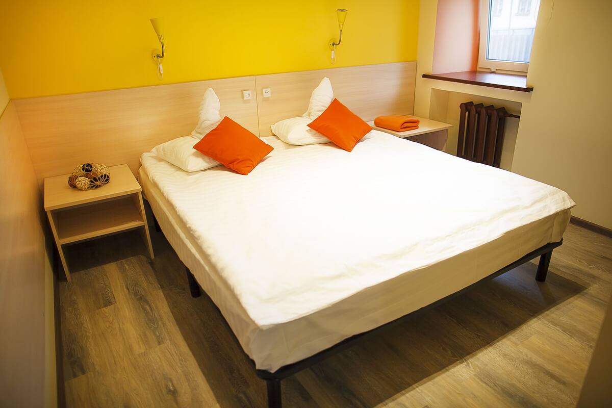 A neatly made bed with white and orange pillows, a wooden headboard, and a bedside table with a decorative item.